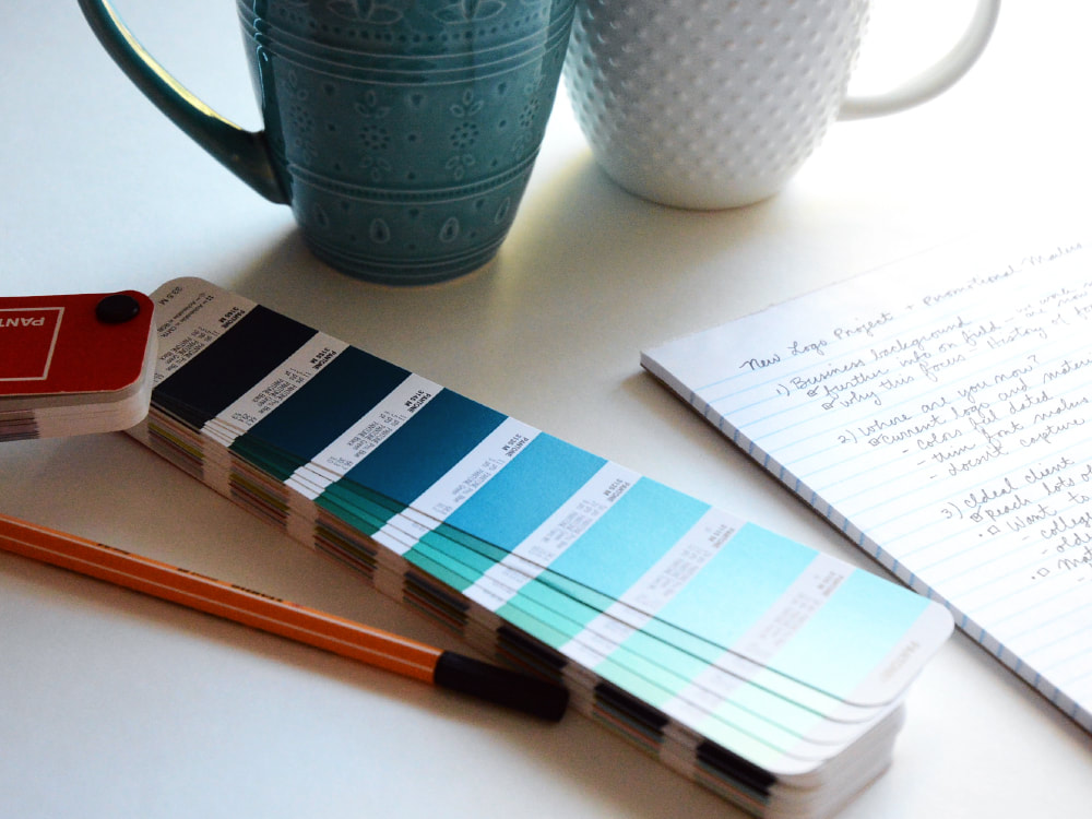 Photograph of a two mugs: a patterned teal one and a white one with raised dots. They sit on a white background behind the teals and greens of Pantone color swatches, a black felt pen, and a notepad with cursive notes about a logo project.