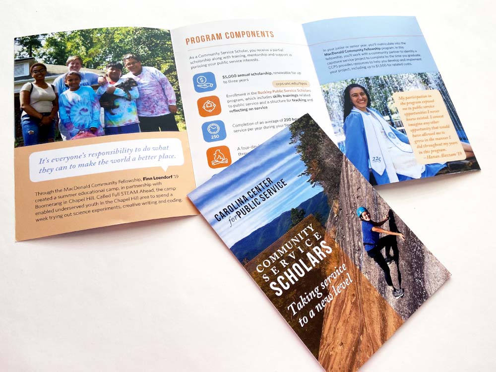 At the top of this photograph is an unfolded trifold brochure. The left panel shows a group of people and a dog with a quote and text below, the center spread has the title “Program Components” followed by various orange and blue icons and text explanations, and the right panel shows a graduating student in her cap and gown sitting at the edge of a fountain, with a quote in front of her. In front of all of this is the cover of the brochure, which shows a college student in a blue shirt and helmet smiling and climbing the side of a mountain. The program logo and title are also on the front of the brochure.