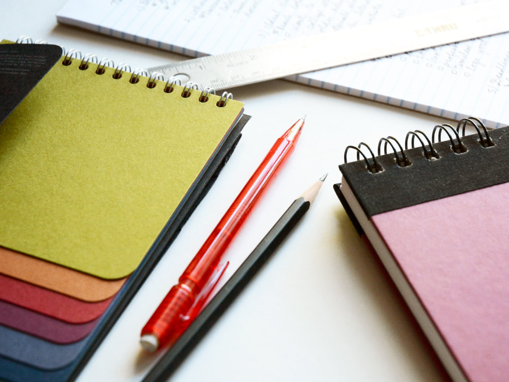 Photograph of a closed maroon and black sketchbook, a drawing pencil, a mechanical pencil, a paper swatch book showing jewel tones, a silver metal ruler, and a lined notepad with cursive notes about a logo project.
