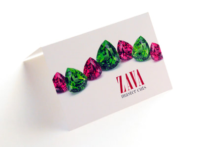 Photograph of a folded white card. On the front is a jaunty curving line of teardrop-shaped pink and green gems, with “Zava Master Cuts” below. On the back is blurry information about gem shows.