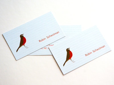Photograph of the fonts and back of a light blue business card with a horizontal column texture on the paper. A a sweet brown and red-orange robin illustration and red-orange text are printed on it.