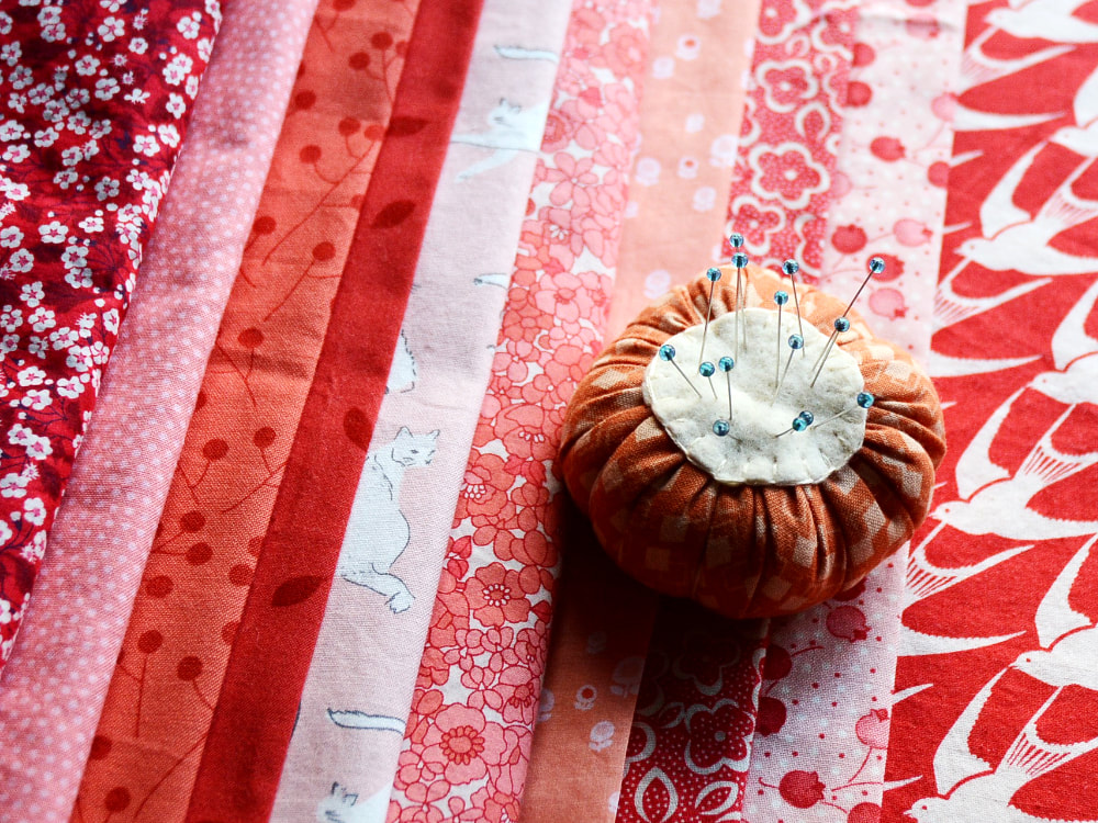 Photograph of a line of folded peach, pink, and red cotton fabrics. Patterns include flowers, dots, leaves, branches, cats, and birds An orange and cream pincushion sits on top and holds pins with blue glass heads.