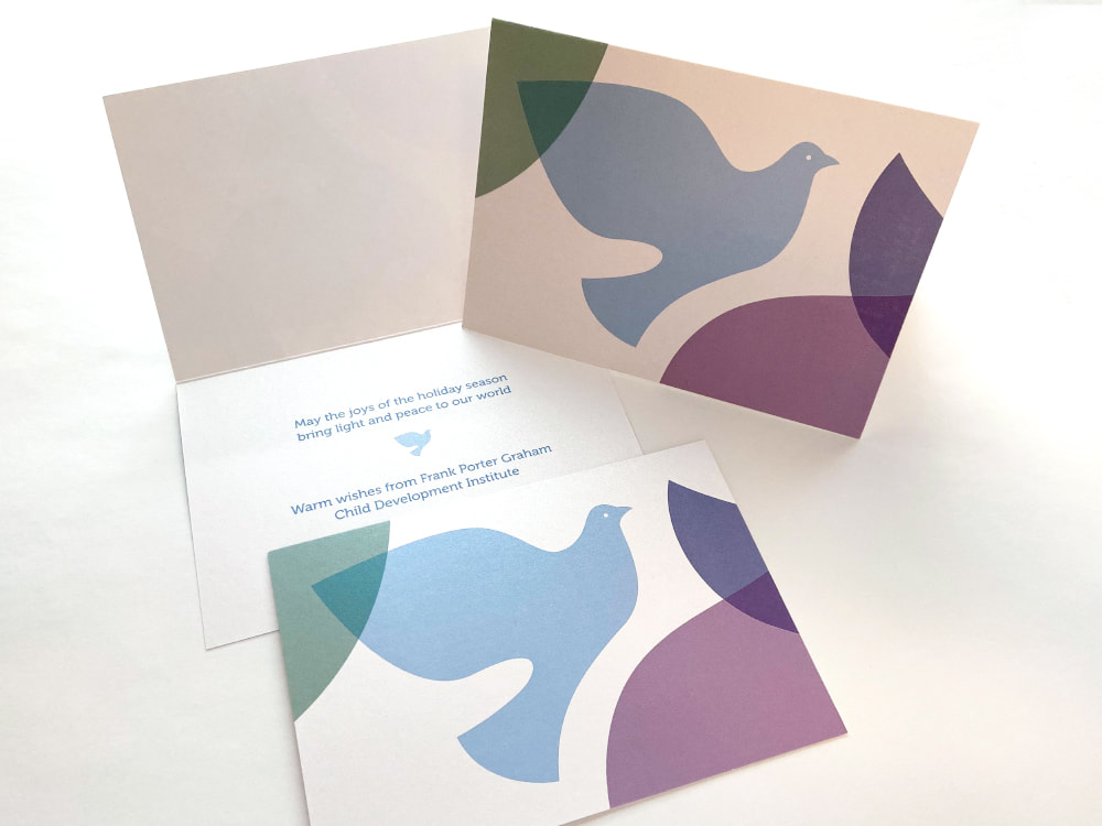 The interior of a holiday card on a pearlized paper reads, in blue ink, “May the joys of the holiday season bring peace and light to our world” with a small blue dove and the organization signature below. The front of the card, shown flat and standing up so you can see the metallic sheen of the ink, shows a larger version of the same Carolina blue dove, overlapping dark green, navy blue, and plum purple leaf shapes. The overlapping areas show transparency, making new colors.